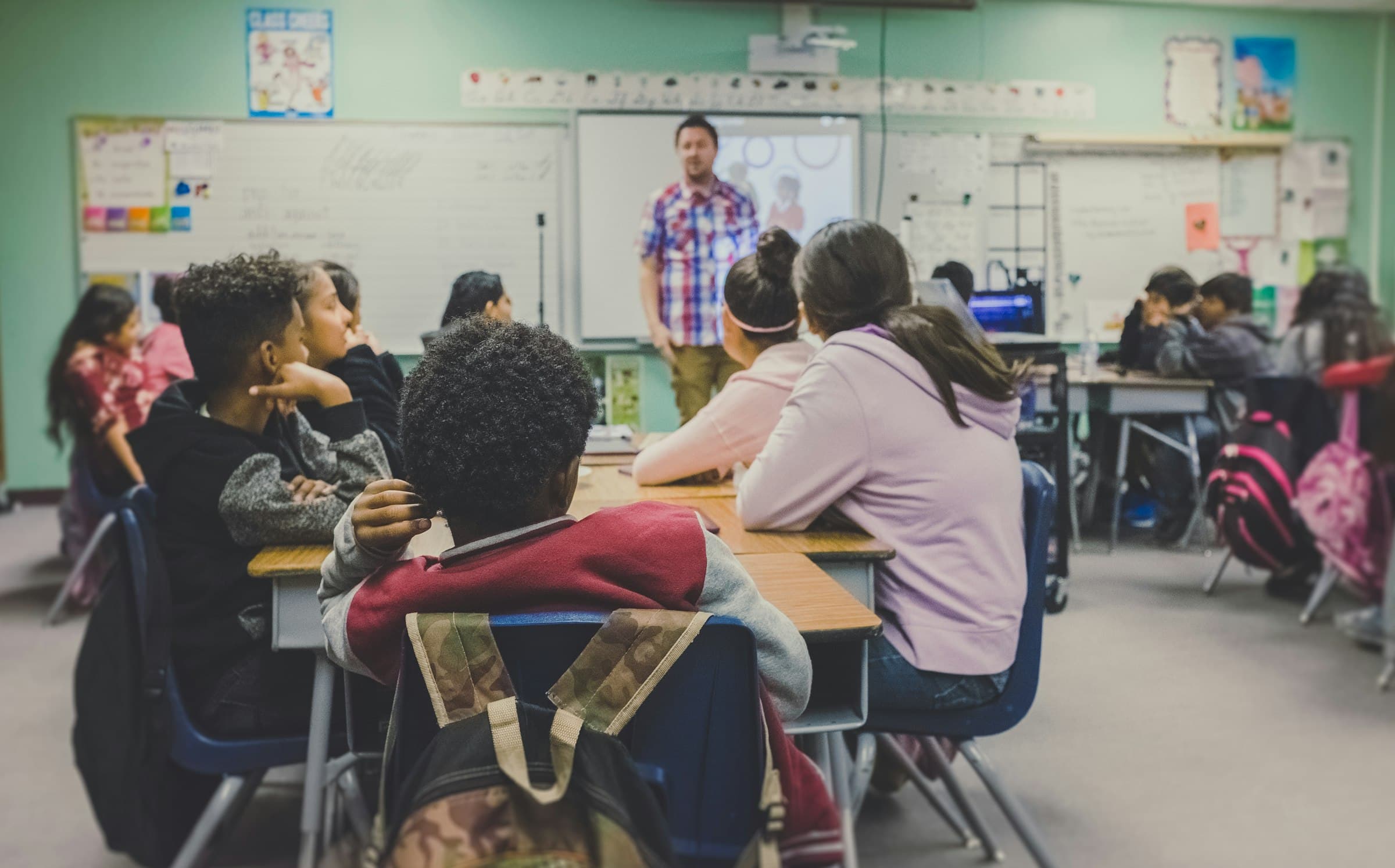 Diverse group of students seated at desks attentively listening to a teacher at the front of a classroom